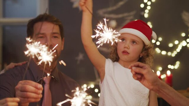 Small girl with parents indoors celebrating Christmas, holding sparklers.