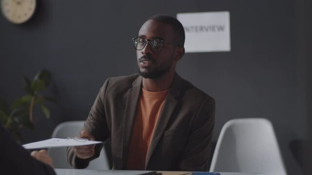 Tracking shot of young African American man sitting at table in the office, giving CV to HR manager and answering questions while having job interview