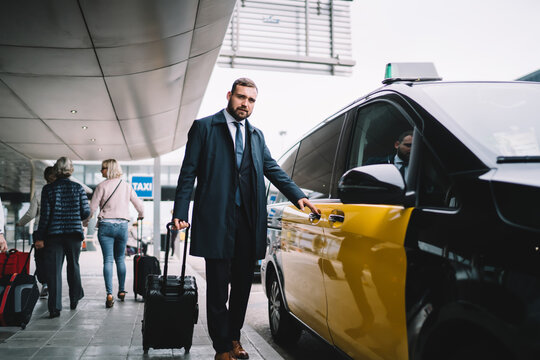 Full length portrait of Caucasian businessman 30 years old dressed in formal clothing posing near taxi transfer from airport, confident male proud CEO with luggage suitcase looking at camera near cab