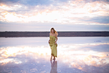 caucasian female with blonde hair in a light green dress on a pink color salty lake with still water against the backdrop of a beautiful pink sunset