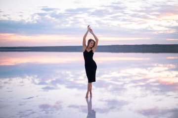 Fototapeta na wymiar beautiful girl with a slim figure in a black dress is staying with raised hands on a salty pink color lake at a vanilla sunset light