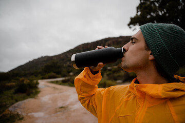 Attractive young male athlete hydrating with water before long distance trail run on mountain path in cold cloudy weather