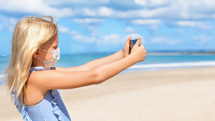 Fototapeta na wymiar Funny girl taking selfie photo by smartphone on tropical sea beach. New rules to wear cloth face covering mask at public places due coronavirus COVID 19. Family holidays with children, summer travel.