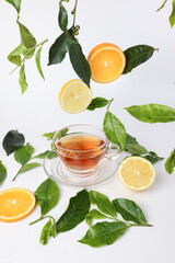 different types of fresh raw green tea leaf flower bud dropping floating elevated lemon orange slice over transparent glass teacup saucer liquid tea on white background