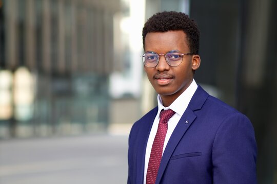 Portrait of young serious black African Afro American businessman standing outdoors business centre, looking at camera in suit, blue jacket, white shirt and tie. 