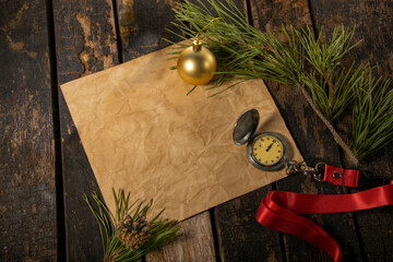 The photo shows an old table on which lies an old sheet of paper with a Christmas tree decoration, a pine twig and a clock.
