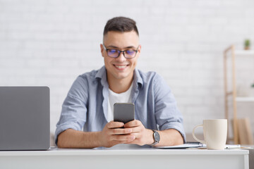 Chat with followers or great reviews and comments. Smiling young guy in glasses typing on smartphone at workplace with laptop in living room interior