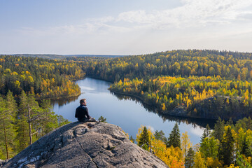 The man on top of a rock in the autumn forest on a background of a beautiful lake. - obrazy, fototapety, plakaty