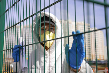 woman in protective mask stands behind fence. holds sheet paper with .drawing virion virus particle. COVID-19. looks at the camera. Coronavirus, health protection, quarantine, safety, pandemic concept
