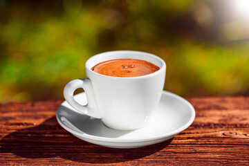 Cup of hot coffee on textured wood board on blurred green nature background outdoors. Natural morning sunlight.