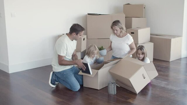 Cheerful family with kids unpacking things in new apartment, sitting on floor with open boxes. Relocation or new home concept