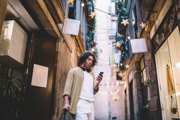 Positive curly male millennial standing on city street with mobile phone browsing web page with locations and destinations, smiling hipster guy holding smartphone reading messages and notifications