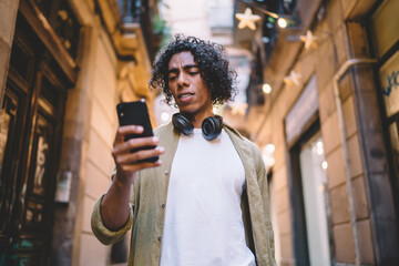 Pensive male hipster reading income message from banking application on mobile phone, serious man millennial checking latest news from websites on smartphone spending free time on city street