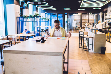 Happy male entrepreneur using modern cellular technology for checking business news connecting to 4g wireless in coworking, successful male freelancer sitting at table desktop and browsing data