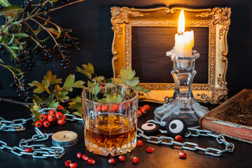 Glass with whiskey on the table next to candles, a book and a branch of kolina on the eve of halloween