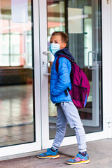 Full length boy wearing protective mask is trying to open the school door. Behind the backpack Schoolboy look at camera