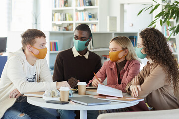 Multi-ethnic group of young people wearing masks while studying together at table in college...