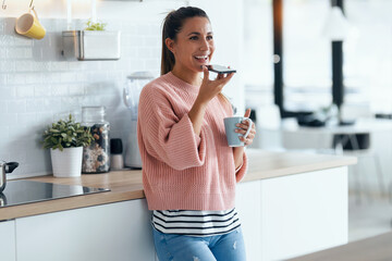 Smiling young woman sending an audio with her mobile phone while drinking a cup of coffee in the kitchen at home.