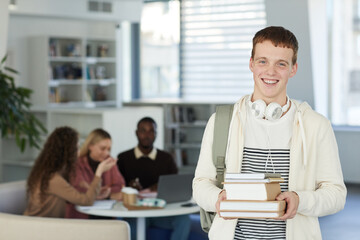 Waist up portrait of smiling teenage boy looking at camera and smiling while standing in college library with people working in background, copy space © Seventyfour