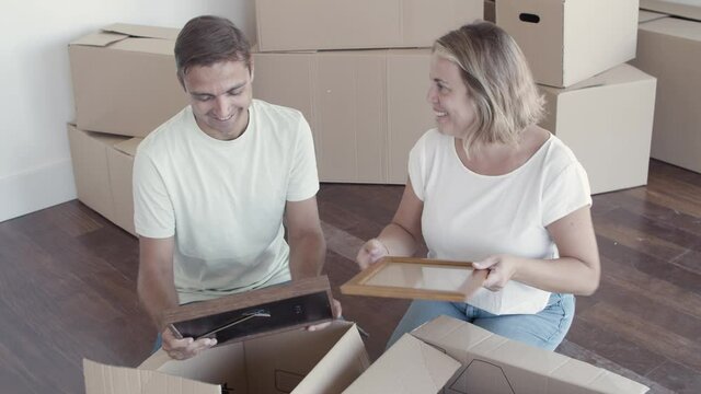 Happy couple talking while packing things for moving, sitting on floor with open boxes. Relocation or new home concept