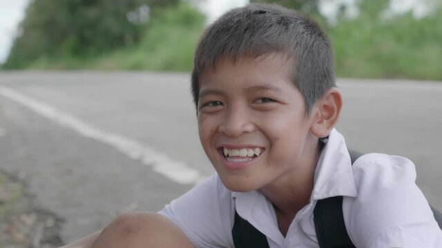 A smiling poor elementary school boy in Thai elementary school uniform is sitting on the ground while going back home after school evening beside the road in the countryside of Thailand. 
