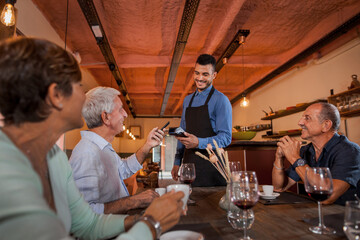 Smiling senior man sitting on table with friends paying with smart phone using NFC technology. Afro american waiter standing and holding a card terminal in a restaurant. Concept paying contactless.
