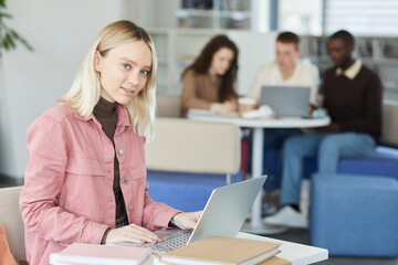 Fototapeta na wymiar Side view portrait of blonde young woman looking at camera while using laptop in college library with people in background, copy space