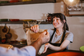 Waitress wearing apron serving customer at counter in restaurant. Young caucasian woman owner offering recycled paper bag with take away food to online client. Small business and service concept.