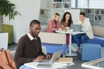 Side view portrait of young African-American man using laptop in college library with people in background, copy space