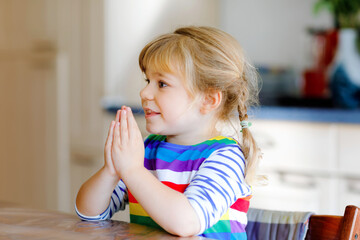 Cute toddler girl praying to God at home. Child using hands for pray and thank for food. Christian tradition.