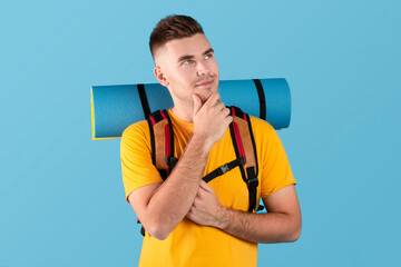 Portrait of attractive young guy with rucksack and tourist mat thinking and touching his chin on blue studio background