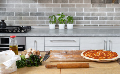 Still life. There are kitchen utensils on the kitchen table, a blank for pizza with tomato sauce, plants in pots in the background.