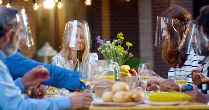 Happy mixed-races family in medical face shields sitting at table with meal outdoors at picnic and talking cheerfully. Joyful young and old people having dinner and speaking at barbrque. Coronavirus.