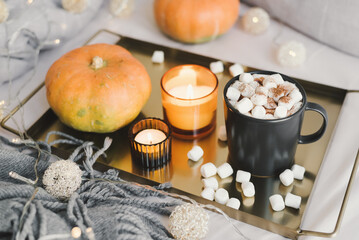 Fototapeta na wymiar Metal tray with hot chocolate and melted marshmallow in a black mug, burning aroma candles and pumpkins served on bed. Fall season inspiration