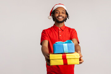 Positive friendly afro-american man with dreadlocks in santa claus hat holding and showing gift boxes, preparing presents for holidays. Indoor studio shot isolated on gray background
