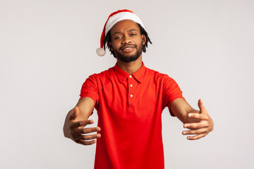 Friendly afro-american bearded man with dreadlocks in santa claus hat holding hands ready to embracing, free hugs on christmas eve. Indoor studio shot isolated on gray background