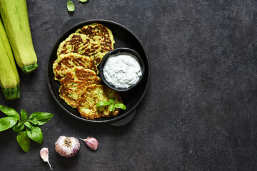 Zucchini pancakes with sauce and garlic in a frying pan on a black background. View from above.