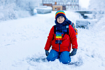 Fototapeta na wymiar Gorgeous little school kid boy with glasses of elementary class walking to school during strong snowfall. Snowy streets in city. Child with backpack or satchel in colorful winter clothes