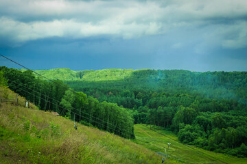 Fototapeta na wymiar Dramatic landscape with thunder sky and green forest. Empty chairlift in ski resort in summer with green grass and no snow.