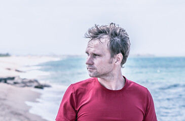Fototapeta premium Serious handsome fair hair fluttering wind man red shirt, stubble face cheeks beard, wrinkled forehead sad, real skin, look far away thinking, blur sea skyline background BANNER. Life problems concept
