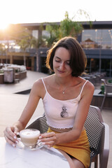 A girl in a yellow skirt and white top sits at a table on the summer terrace of a cafe and looks on the cup of coffee. On vacation in Turkey hotel. Smile. Sunny day. Morning. On the sea.