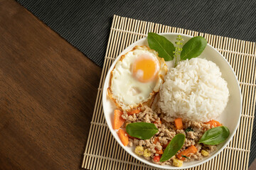 Top view, Stir-fried Thai basil with minced pork meat, carrots, and baby corns, with fried egg and fresh basils sort in a white dish on bamboo placemat, grey placemat, brown wood table. Copy space