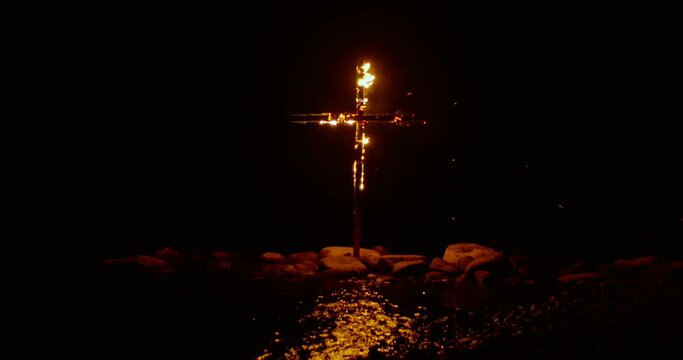 Mystical lockdown shot of wooden cross burning in sea water surrounded by stones at dark night, sparks flying away from flame
