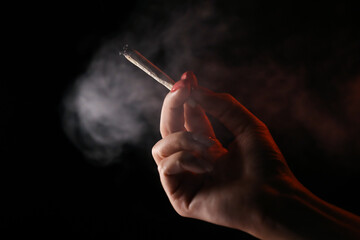 Woman hand holding cigarette in a smoke against black background. Smoking cannabis joint. Medical...