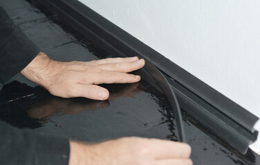 A man is installing a black PVC skirting board along a white wallpapered wall and hardwood painted...