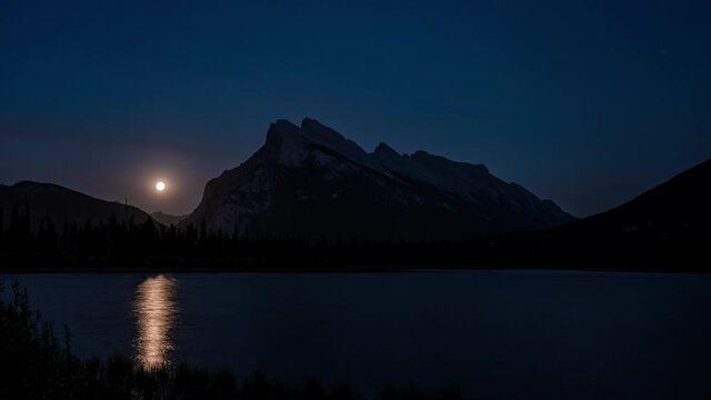 Mount Rundle with moonrise in summer night. View from Vermilion Lakes lakeshore. Bright full moon light up the lake, golden reflection on surface. Banff National Park, Alberta, Canada. Time-lapse 4K