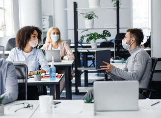 Team of employees working on project and social distancing during outbreak of covid-19. Millennial male manager in protective mask talk to european and african american women