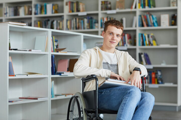 Portrait of smiling young man using wheelchair in school library and looking at camera, copy space