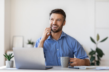 Cheerful male entrepreneur talking on mobile phone and drinking coffee in office