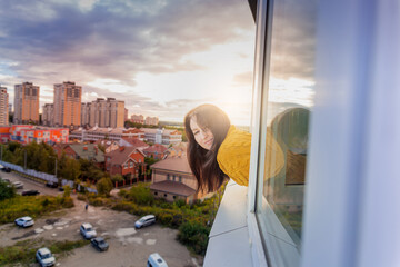 A young beautiful girl in a yellow sweater looks out of the window on the eighth floor against the backdrop of the sunset and smiles.